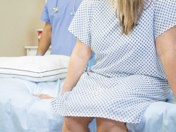 Woman in patterned hospital gown sitting on bed with healthcare professional in blue scrubs