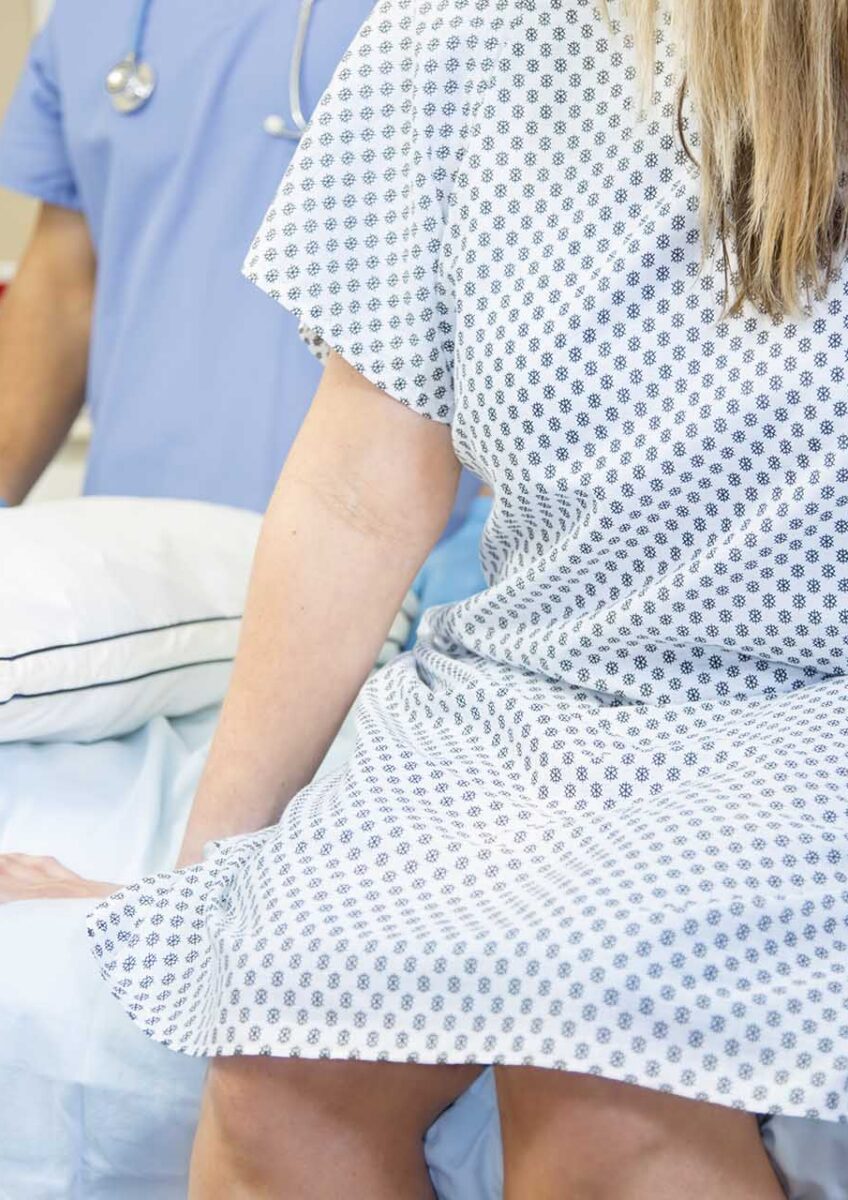 Woman in patterned hospital gown sitting on bed with healthcare professional in blue scrubs